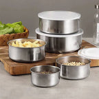 A set of stainless steel food storage containers with white plastic lids, shown in different sizes with food items like peanuts, seeds, and chopped vegetables inside. The containers are arranged on a wooden cutting board on a gray countertop.